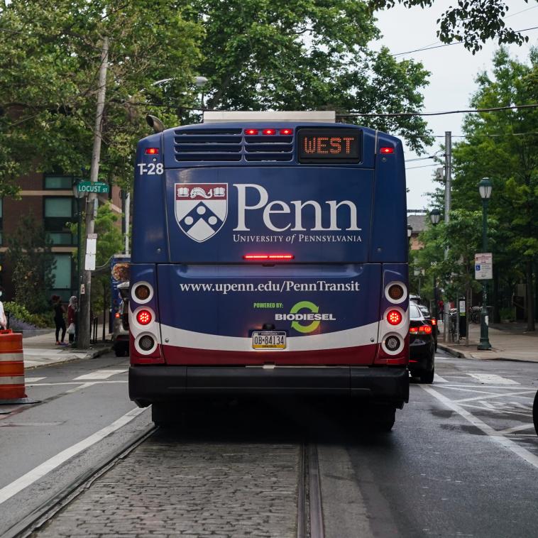 A bus is seen waiting at a stop sign at an intersection on Locust street. The perspective is taken from a car waiting behind the bus. The bus has a wrapper advert with a large Penn emblem on the back, and below that it reads “University of Pennsylvania” and in a larger font: “www.upenn.edu/PennTransit”. The bus also advertises "Powered by BIODIESEL" which appears above the bus license plate. 
