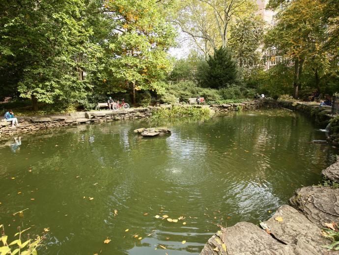 a lake feature in an urban park. Students are sitting on stones and benches lining the lake. On the far end a couple can be seen sitting at a picnic table. 