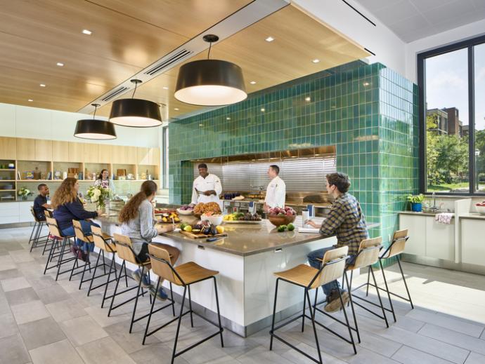 Quaker kitchen at Gutmann college house is shown. It has a large kitchen island with 10 stools around it. On the island, there are several trays of food. Several students are seated around the table. Two people wearing chef uniforms are standing behind the island at the stoves and giving a cooking demonstration.