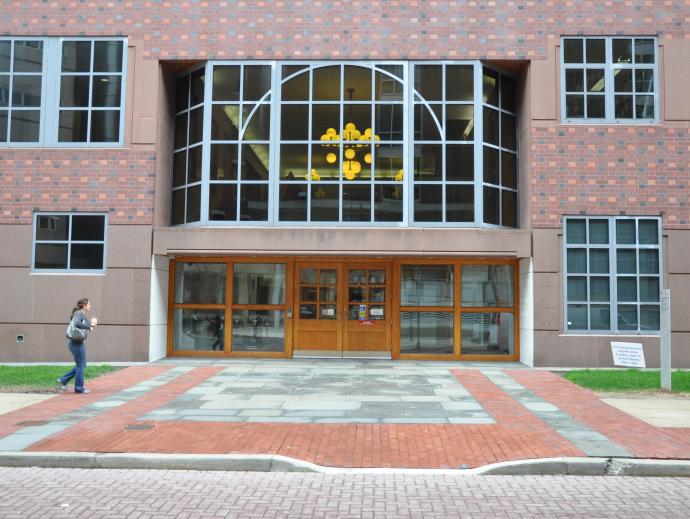 The entryway and doors of a building are shown. A woman holding a soft drink is walking by the pathway leading to the doors.