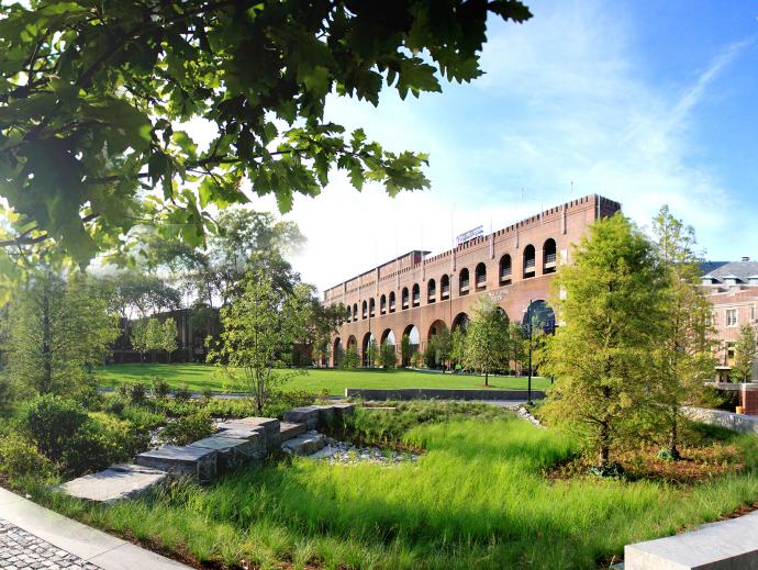 a welcoming open space of lawn which is surrounded by tree-lined walkways. A flagpole with sculpted figures as the base stands on a pillar of steps along one of the walkways. 