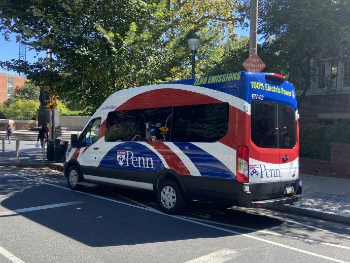 A van-like minibus is seen parked by the side of the street. It is covered with red white and blue ribbon artwork and has the Penn emblem on it. At the top of the bus there are green letters that read “100% Electric Power” and “ZERO EMISSIONS”.