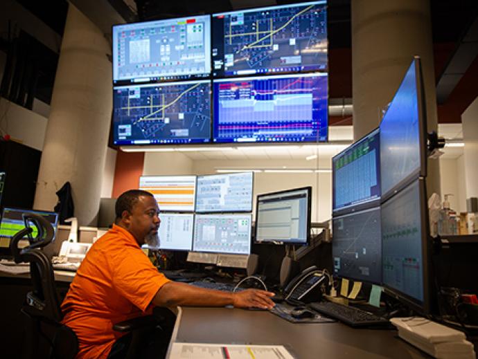 man in orange shirt at desk with many computer monitors