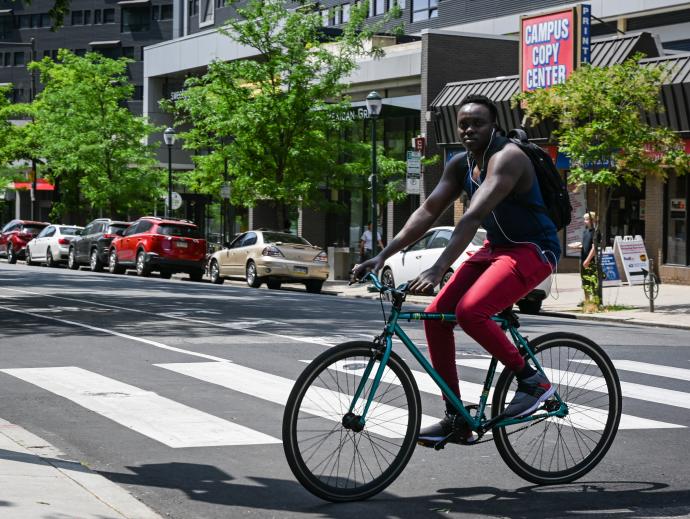 A woman is seen riding a bicycle while crossing a street near a crosswalk. Behind her, a shop has a sign that reads Campus Copy Center.