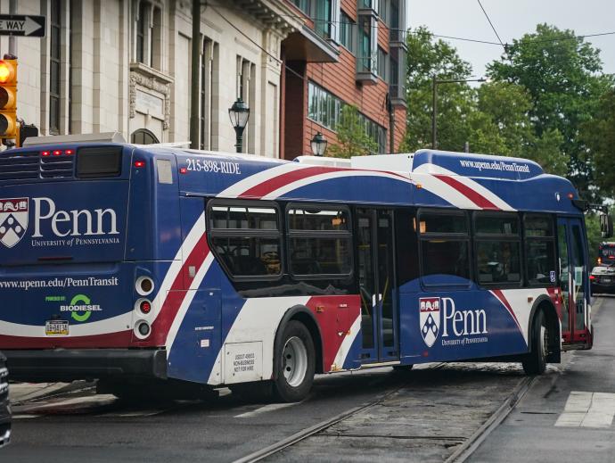The picture is taken from the perspective of someone waiting at a stop light  - at the corner ahead, a bus is seen turning onto the street. The bus has a wrapper advert covering the outside with red white and blue ribbon artwork, with a large Penn emblem and “University of Pennsylvania”  decorating both the back and the side of the bus. On the back it also says “www.upenn.edu/PennTransit” and "Powered by BIODIESEL” 